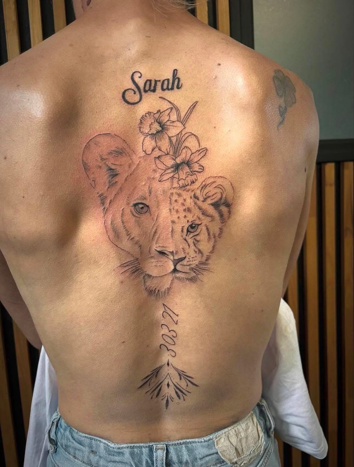 Tatouage fine line et micro-réalisme : portrait de lionne avec prénom Sarah, jonquilles et date sur le dos