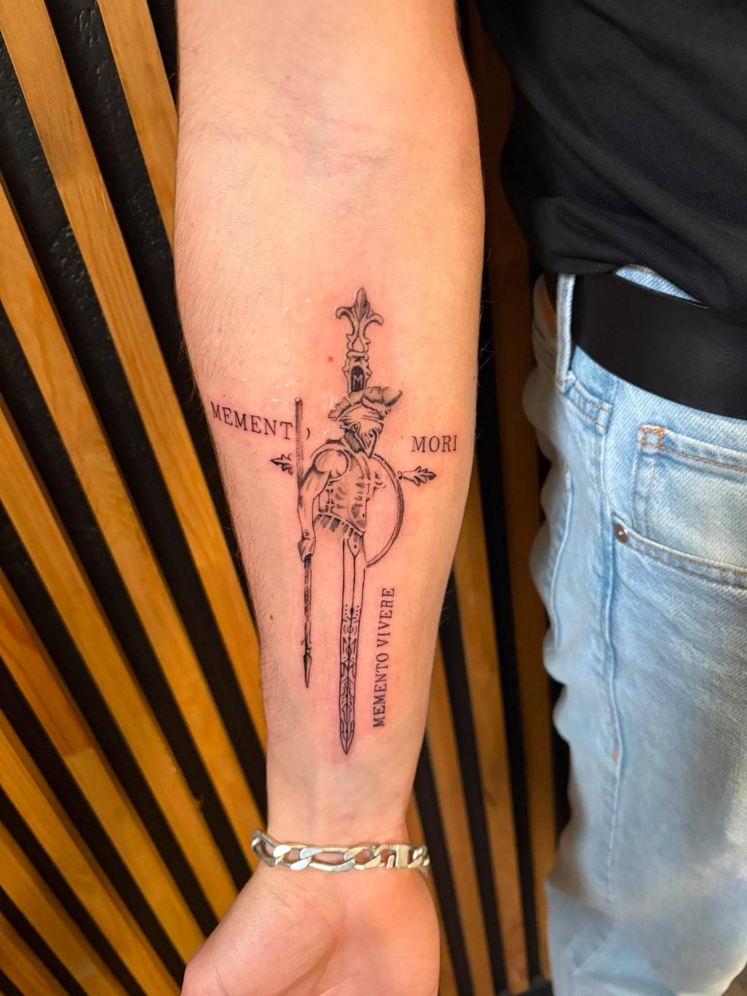 Tatouage fine line et micro-réalisme : guerrier spartiate, épée et inscriptions Memento Mori sur l’avant-bras