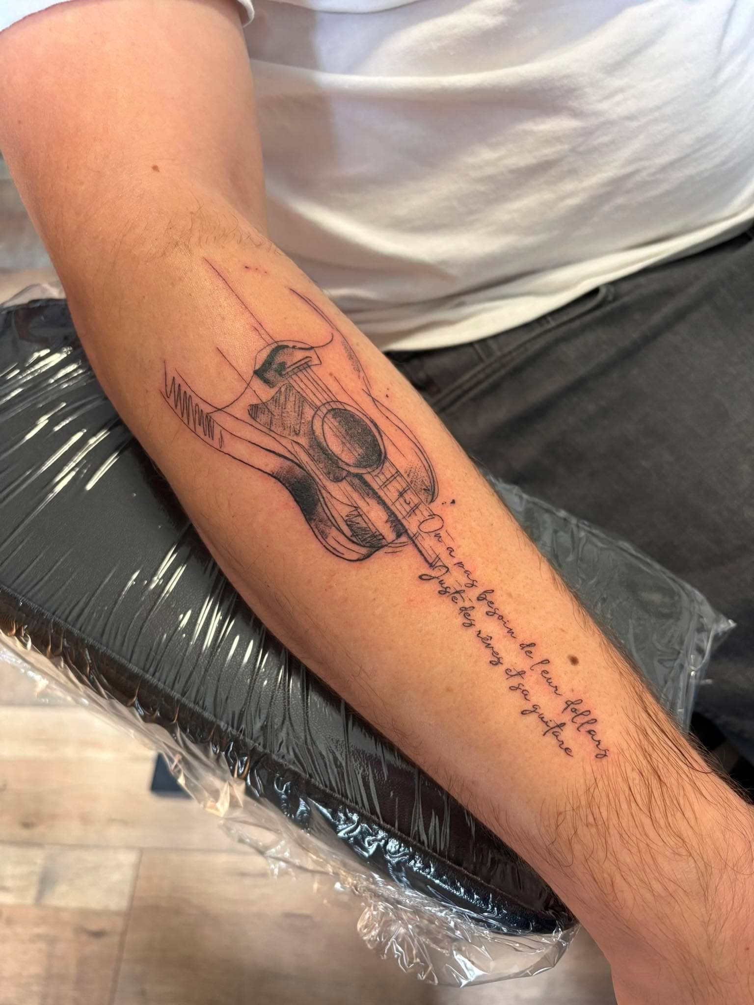 Tatouage fine line : guitare esquissée avec citation en script sur l’avant-bras